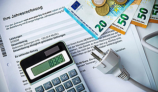 Eine Jahresverbrauchsabrechnung liegt auf einem Tisch. Darauf befinden sich ein Taschenrechner mit angezeigtem Betrag, Euro-Scheine und -Münzen sowie ein weißer Netzstecker, die gemeinsam Kosten und Energieverbrauch symbolisieren.