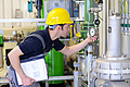 Ein Anlagenmechaniker mit gelbem Schutzhelm prüft in einer Industrieanlage ein Manometer an einer Rohrleitung und hält dabei ein Klemmbrett mit Unterlagen in der Hand.